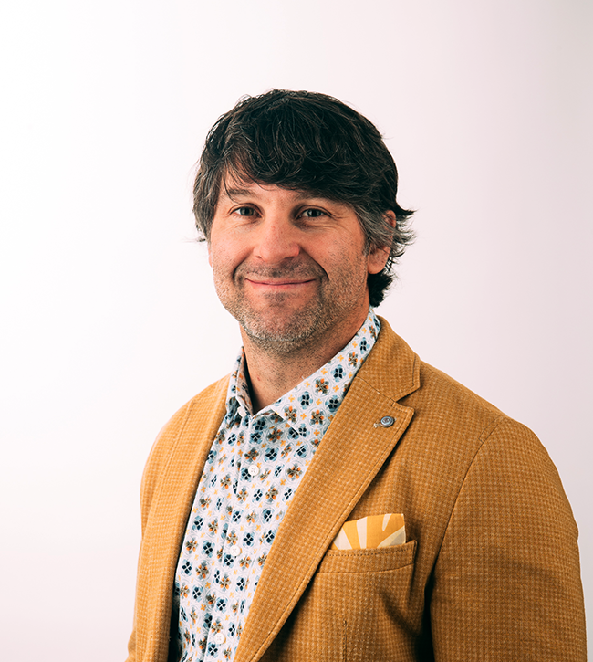 Professional headshot of a smiling man with dark hair, wearing a mustard-coloured blazer, patterned button-up shirt, and pocket square on a neutral background.