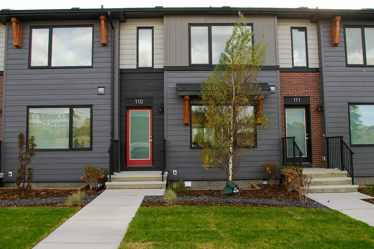 A modern residential development featuring multi-story townhouses with charcoal-gray and white siding, red brick accents, and natural wood corbels.