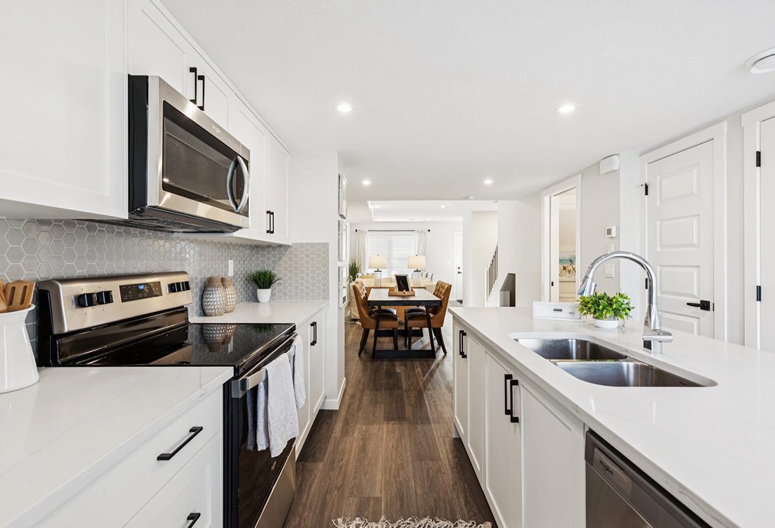 A bright, modern open-concept kitchen featuring white cabinetry, stainless steel appliances, and a view into a dining area with a dark wood table and tan leather chairs.
