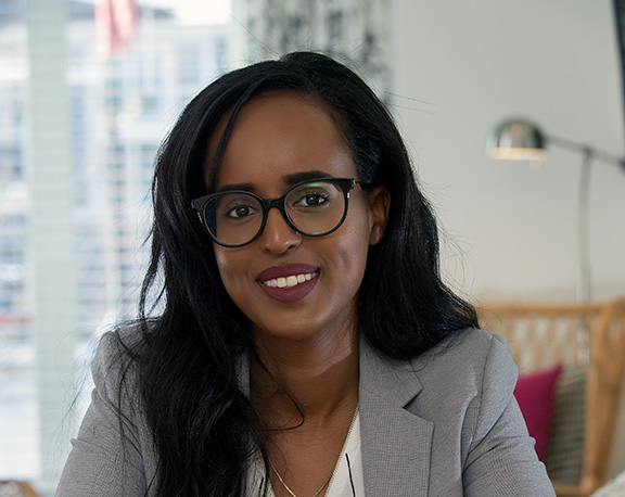 A professional headshot of a smiling woman with long dark hair and black-framed glasses, wearing a grey blazer over a white blouse.