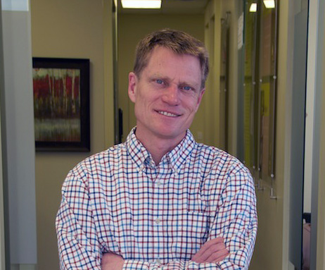 A headshot of a smiling man with short light-brown hair, wearing a red, white, and blue checkered button-down shirt, standing with his arms crossed in an office hallway.
