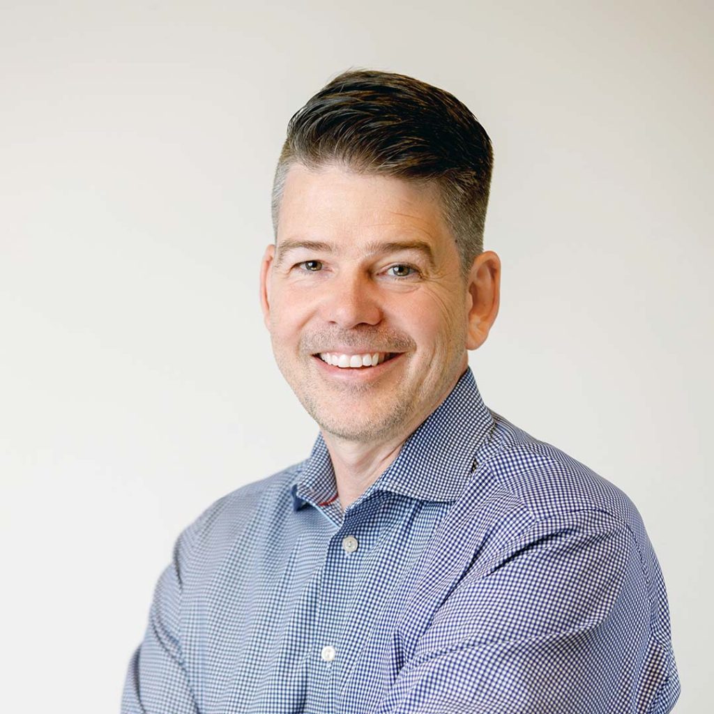Professional portrait of a man with a warm smile and short hair. He is wearing a blue checkered dress shirt and is positioned against a clean, neutral white background.