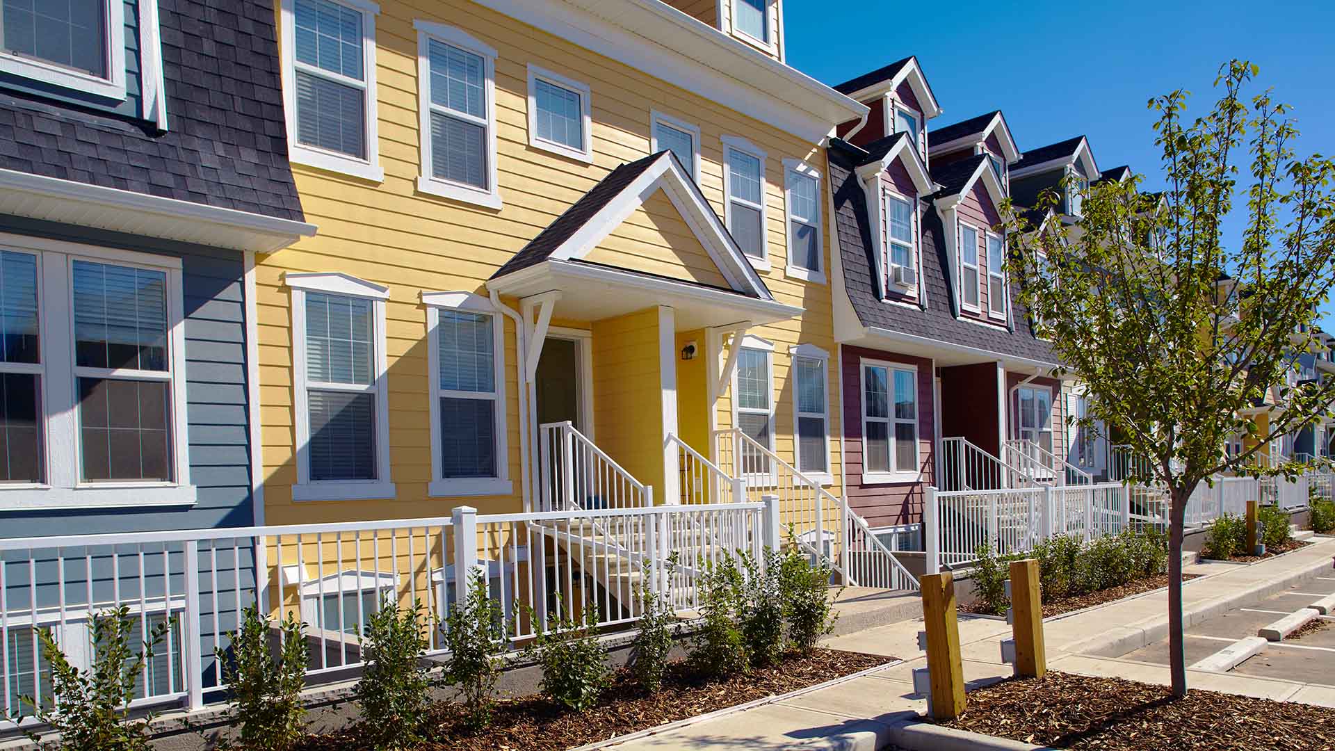 A multi coloured two-storey townhome building with a white metal fence, grey concrete sidewalk and green shrubs and a tree in front
