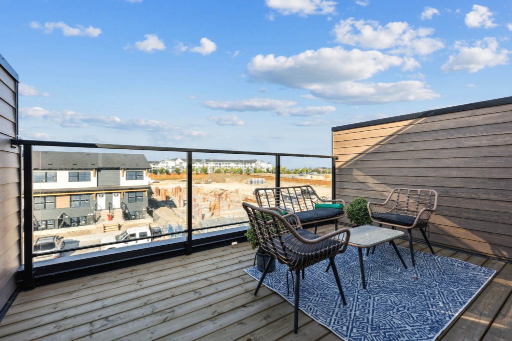 A rooftop patio staged with modern woven patio furniture and a patterned rug. The balcony features a privacy wall and black railings, overlooking a neighborhood of modern townhomes under a blue sky with light clouds.