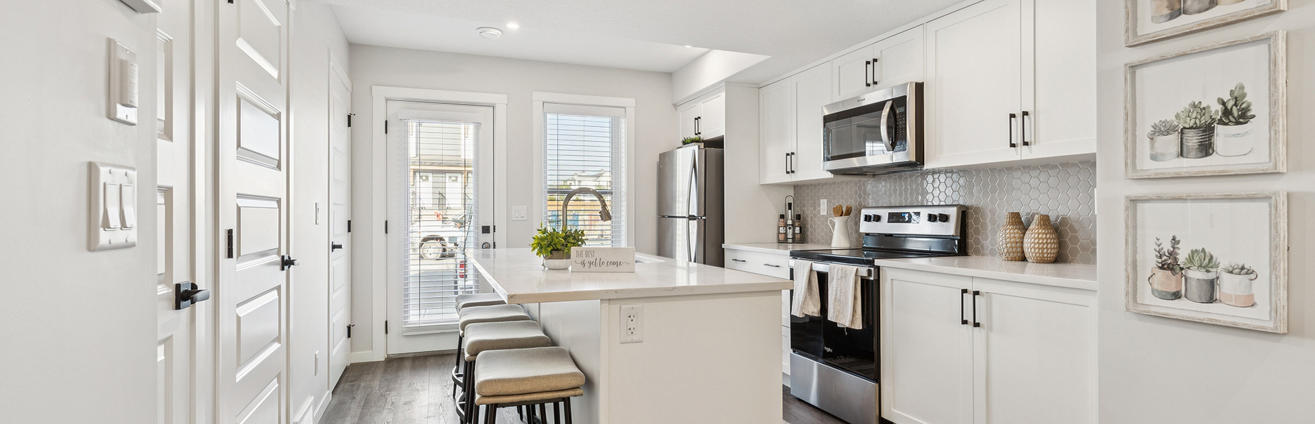 A wide-angle view of a modern apartment interior featuring a white kitchen with a large island and breakfast bar. To the left, a hallway with several white doors leads toward the backyard, while the kitchen transitions into a bright living area.