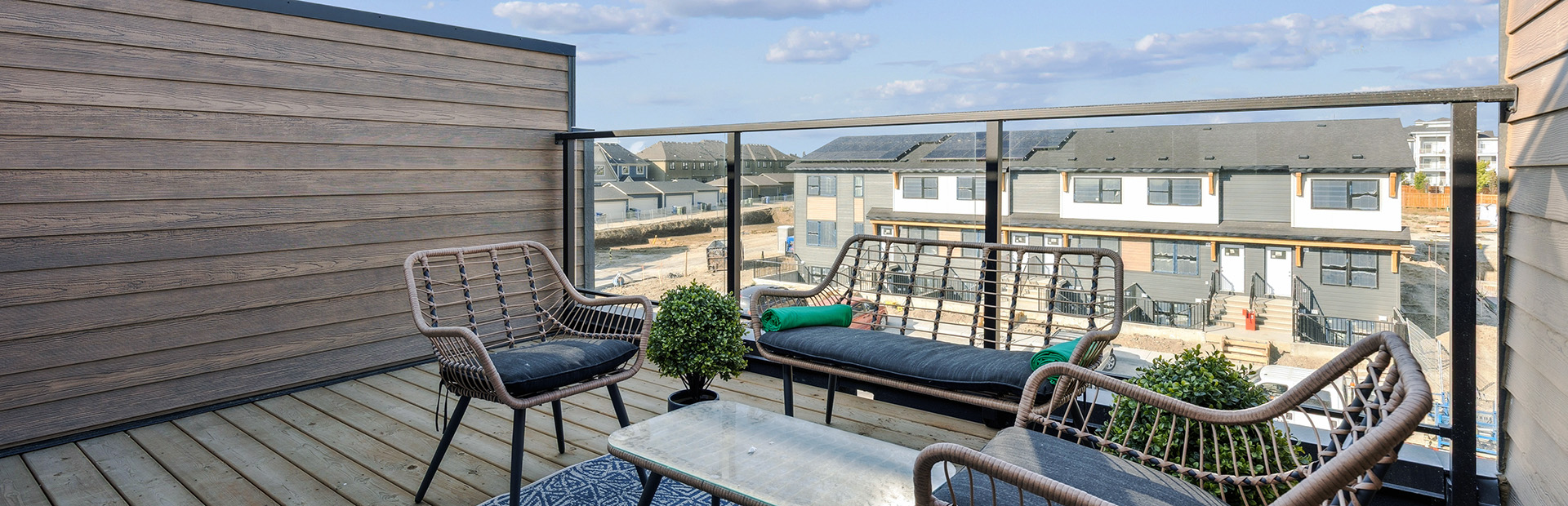 A rooftop patio staged with modern woven patio furniture and a patterned rug. The balcony features a privacy wall and black railings, overlooking a neighborhood of modern townhomes under a blue sky with light clouds.