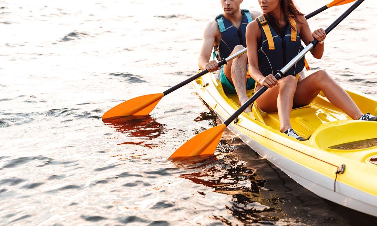 Couple paddling a yellow kayak together on a calm lake at sunset, wearing life jackets.