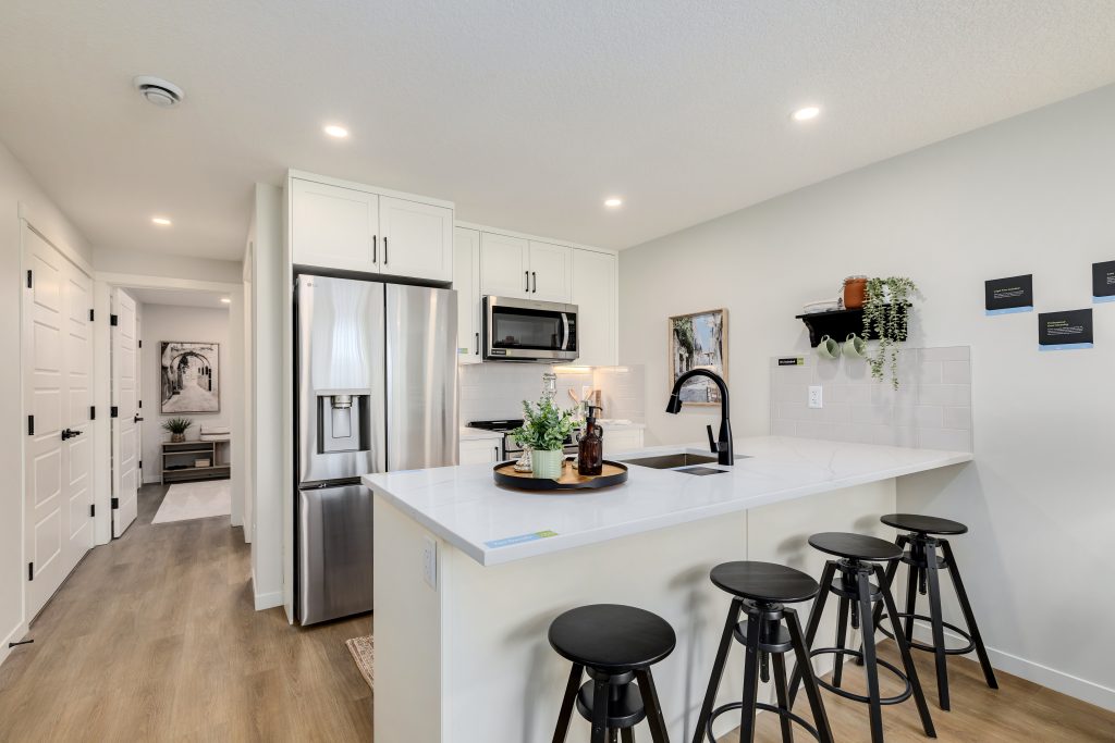 Open-concept modern kitchen featuring white cabinets, stainless steel refrigerator and microwave, quartz island with black bar stools, and light wood flooring.