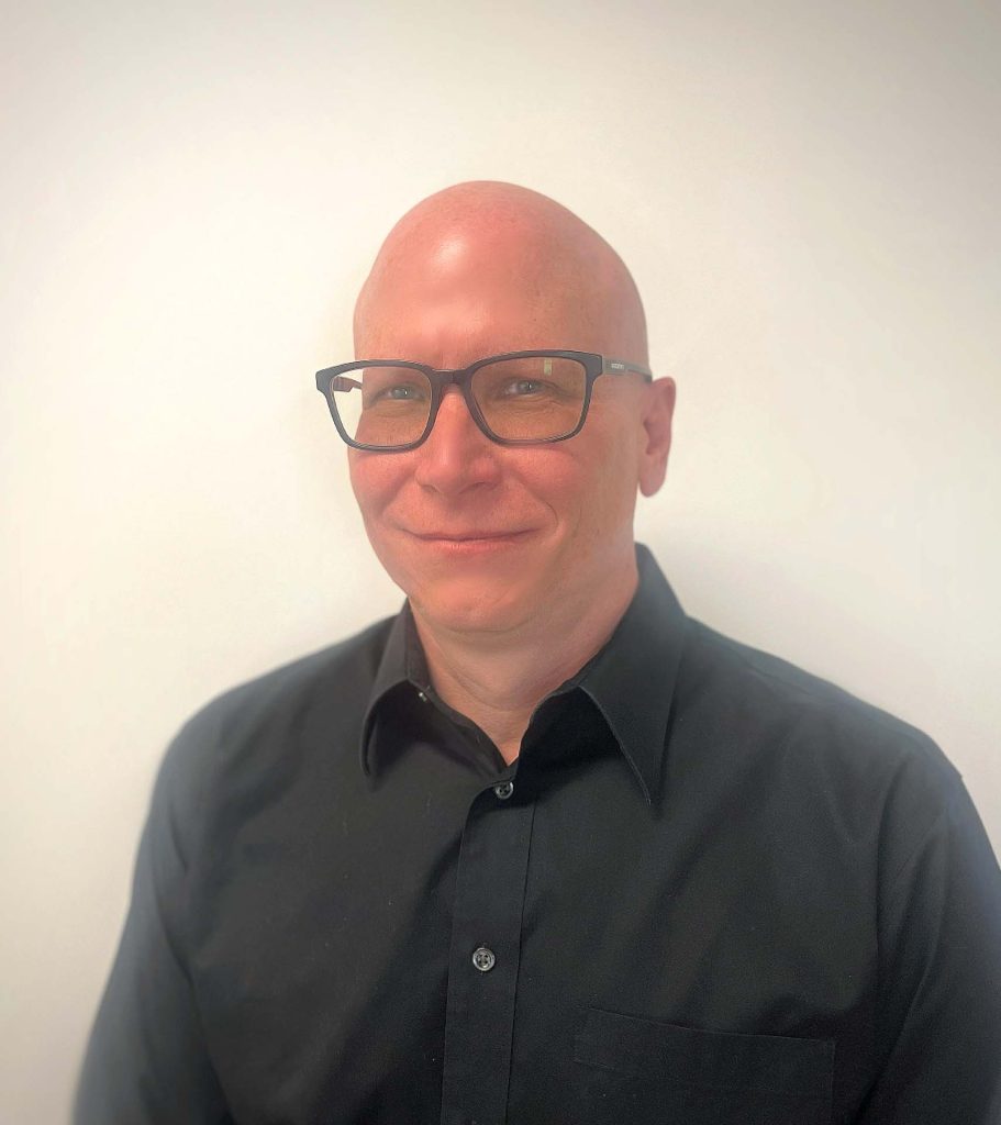 A headshot of a smiling man with a shaved head and black-rimmed glasses, wearing a black button-down shirt against a plain white background.