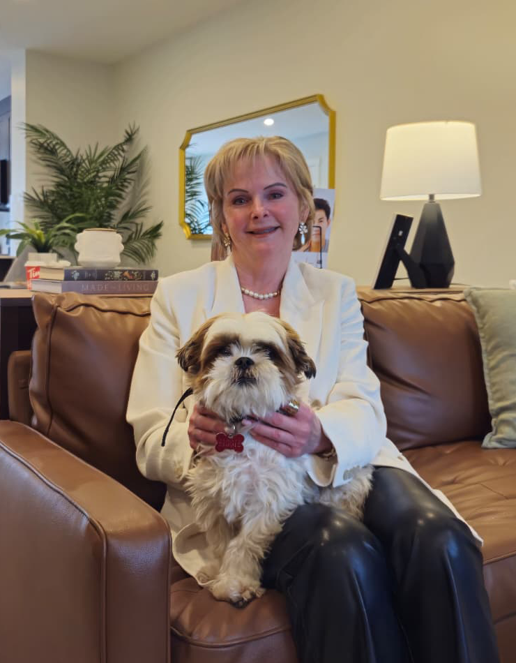 A smiling woman with blonde hair wearing a white blazer and pearl necklace, sitting on a brown leather sofa and holding a small brown and white Shih Tzu dog.