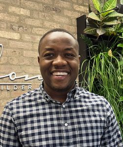 Team member George Daniel smiling in front of Avalon Homes signage and greenery wall, wearing checkered shirt.