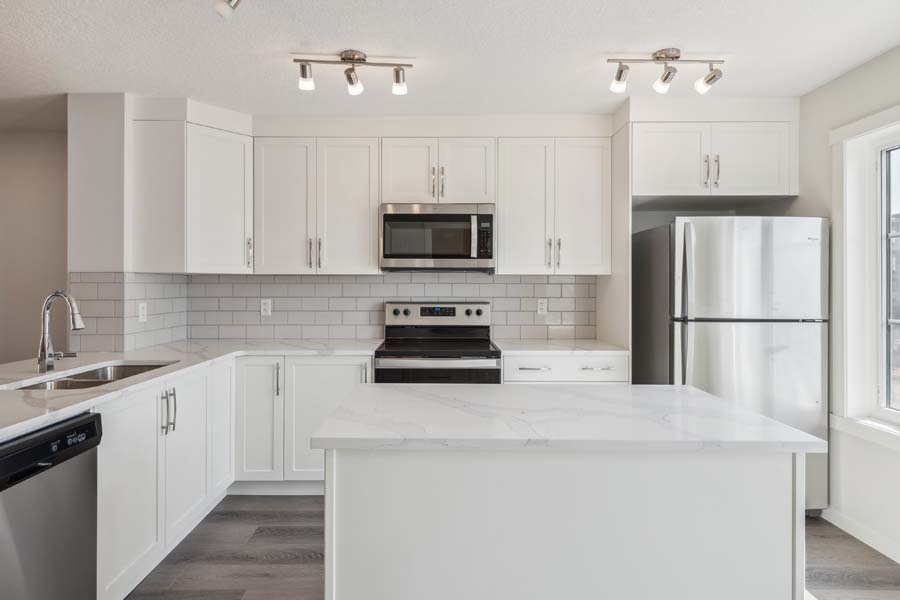 A bright, modern kitchen with white Shaker cabinets, light marble-patterned quartz countertops, and a subway tile backsplash. The space includes a large kitchen island, stainless steel appliances, and dark wood-grain flooring.