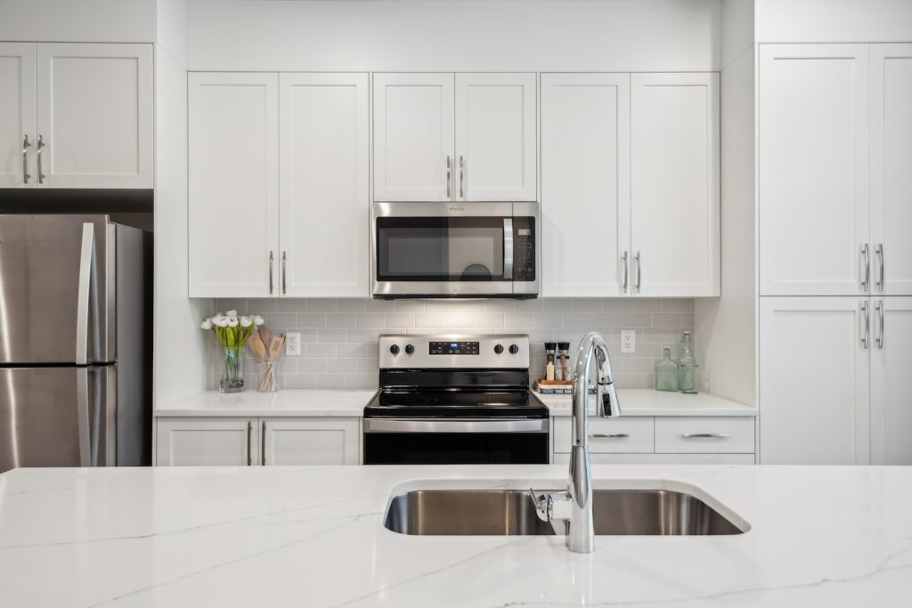 Open-concept modern kitchen featuring white cabinets, stainless steel refrigerator and microwave, quartz island with black bar stools, and light wood flooring.