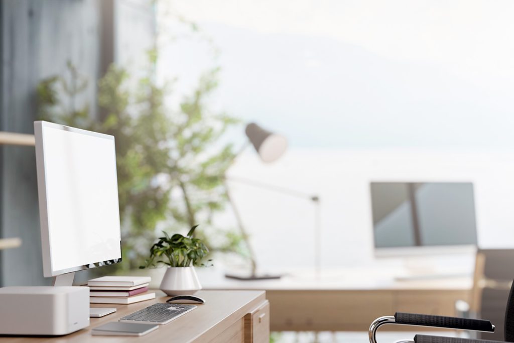A minimalist home office with a light wood desk featuring a modern silver computer, a small potted plant, and a stack of books. The background is brightly lit and softly blurred, showing a secondary workstation and greenery.