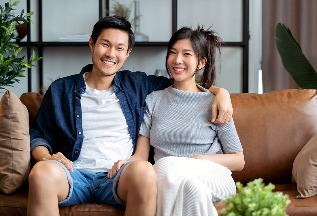 A smiling young couple sitting together on a brown leather sofa in a bright, modern living room.
