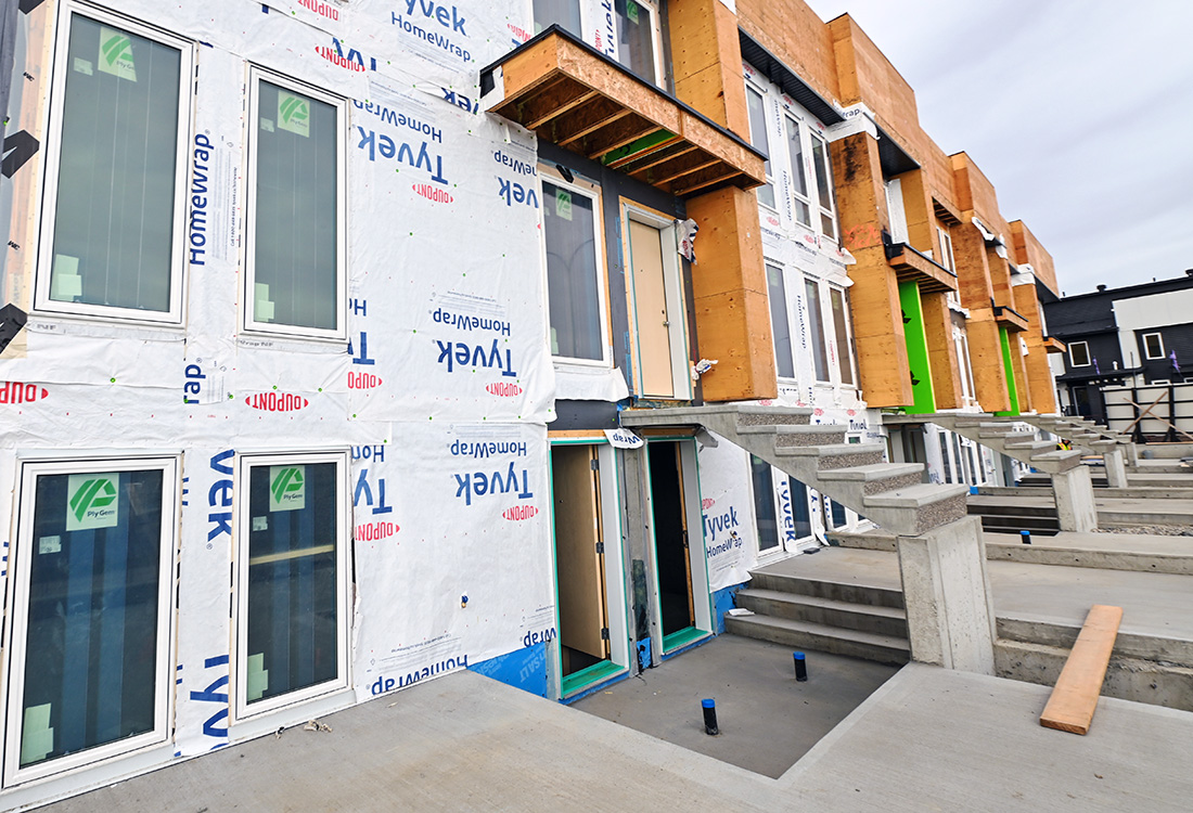 Multi-unit residential townhomes under construction featuring Tyvek house wrap, installed windows, and poured concrete exterior stairs.
