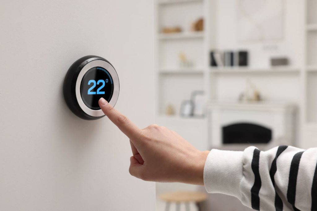 A close-up shot of a person's hand adjusting a sleek, round smart thermostat on a white wall, displaying a temperature of 22 degrees.