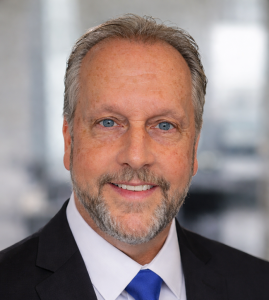 Gemini said A professional headshot of a middle-aged man with blue eyes, a light complexion, and short, salt-and-pepper hair. He has a neatly trimmed grey and brown beard and mustache, and is smiling at the camera. He is wearing a dark suit jacket, a white dress shirt, and a bright blue necktie against a blurred office background.