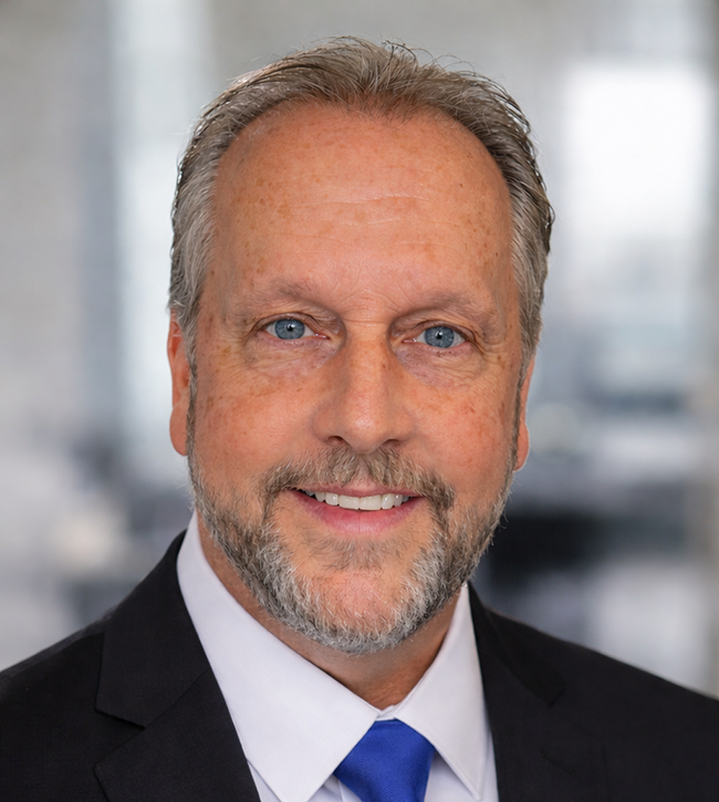 Gemini said A professional headshot of a middle-aged man with blue eyes, a light complexion, and short, salt-and-pepper hair. He has a neatly trimmed grey and brown beard and mustache, and is smiling at the camera. He is wearing a dark suit jacket, a white dress shirt, and a bright blue necktie against a blurred office background.